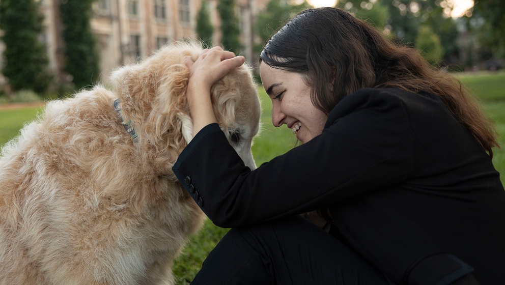 A smiling student in a dark blazer leans forward to pet a fluffy golden retriever assistance dog on a grassy campus lawn, showcasing a joyful moment of connection.