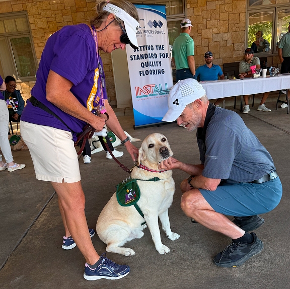 A woman wearing a purple CHAMP shirt stands next to a yellow Labrador retriever service dog wearing a green vest as a crouched man wearing a white cap gently pets the dog during an outdoor community event.