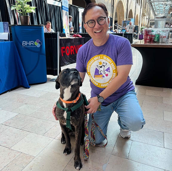 A smiling man wearing a purple CHAMP Assistance Dogs t-shirt kneels next to a brindle dog wearing a green harness inside a large indoor convention or community event hall.