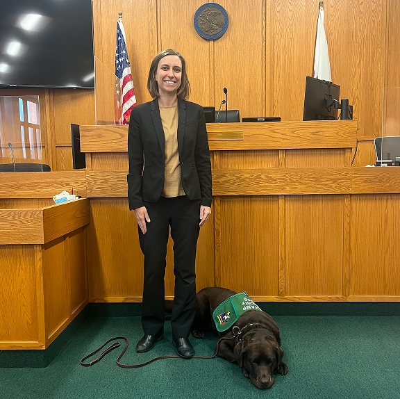 A woman in a black pantsuit stands in a wood-paneled courtroom alongside a chocolate Labrador retriever facility dog lying down calmly at her feet while wearing a green CHAMP vest.
