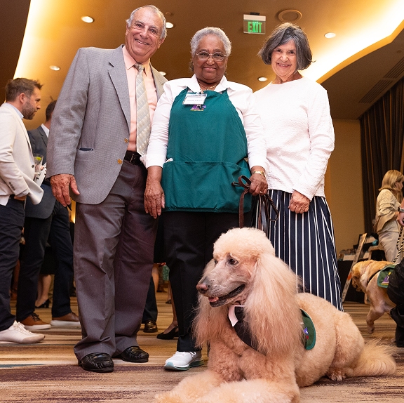 Three smiling individuals pose together at a formal gala event with a large, light-colored standard poodle wearing a CHAMP Assistance Dogs vest lying down calmly in the foreground.