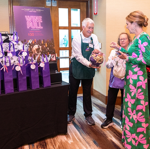 A volunteer wearing a green CHAMP apron holds a glass bowl filled with wine corks for a guest at a "Wine Pull" fundraising station during the Champ's Best Friend Gala.