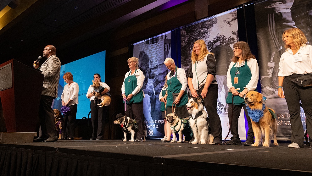 A speaker addresses an audience from a podium on a stage while a line of CHAMP Assistance Dogs volunteers and handlers stand proudly with their dogs during a formal presentation event.