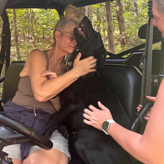 A woman sitting behind the wheel of an outdoor utility task vehicle (UTV) smiles warmly and hugs a happy black Labrador retriever that is leaning in to cuddle her.