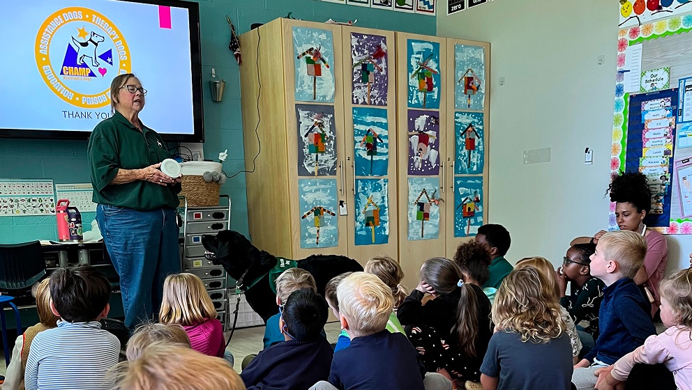 A presenter in a green shirt stands before an attentive classroom of young children, speaking next to a black assistance dog and a digital screen displaying the CHAMP Assistance Dogs logo.