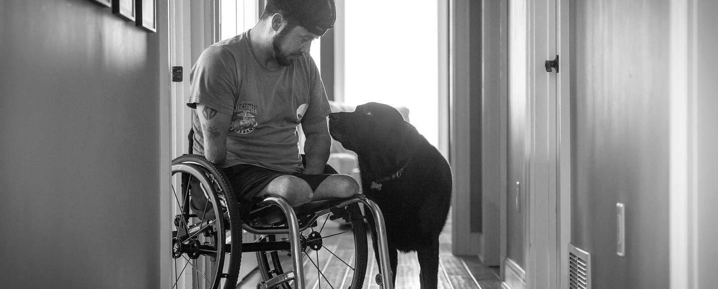 A black and white photograph of a man using a wheelchair looking down affectionately at a black Labrador retriever service dog standing by his side in a home hallway.