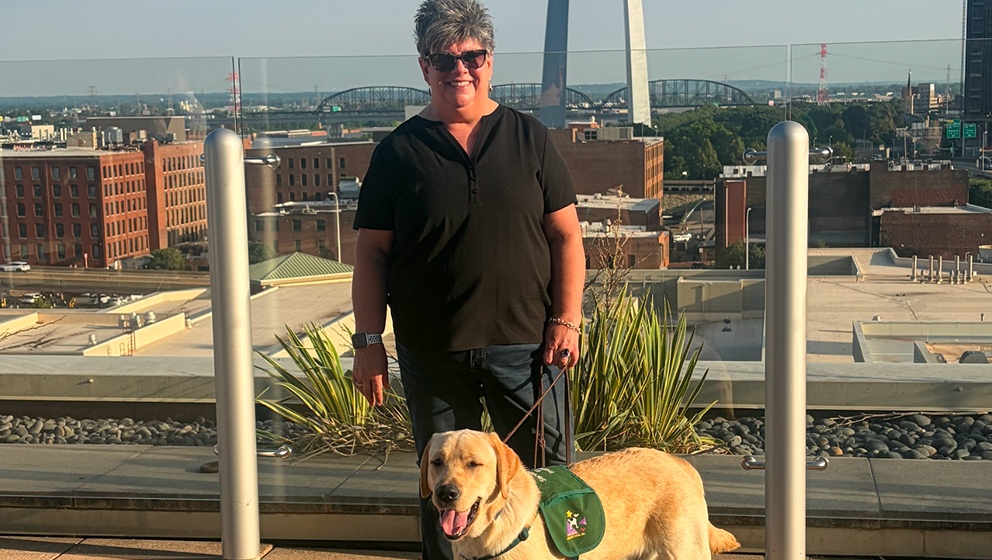 A smiling woman stands on an outdoor rooftop terrace with a yellow Labrador retriever wearing a green CHAMP vest, with the St. Louis Gateway Arch visible in the background skyline.