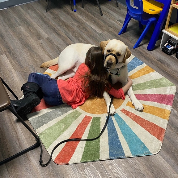 A young girl in a coral shirt lying on a colorful sunburst rug and warmly hugging a yellow Labrador retriever CHAMP assistance dog wearing a black head halter.