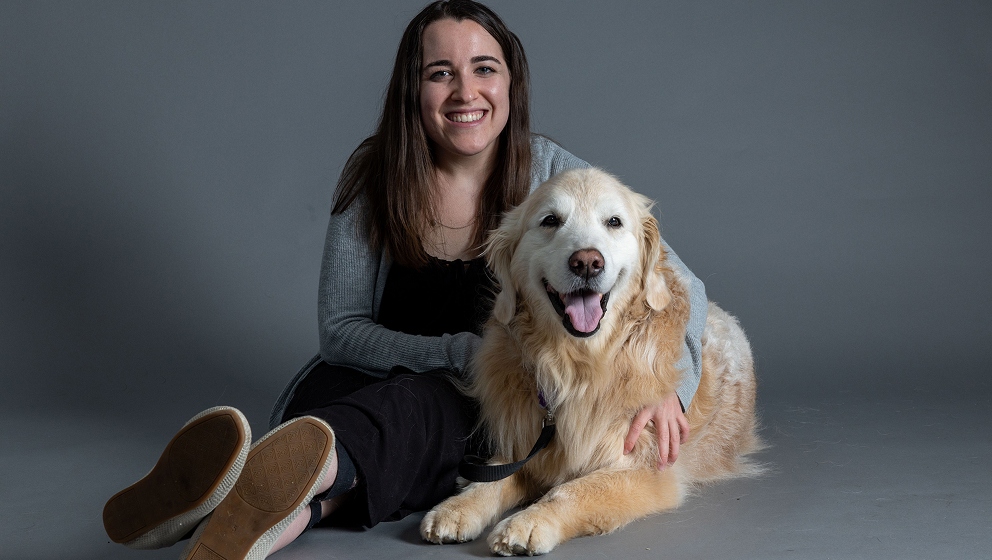 A close-up, front-facing portrait of a golden-colored yellow Labrador retriever service dog looking directly into the camera with an attentive and gentle expression sitting next to a young woman with brown hair.