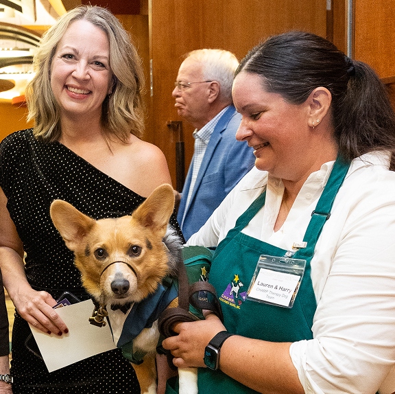 A smiling CHAMP therapy dog team volunteer, wearing a green apron with a name tag that reads "Lauren & Harry," holds a Pembroke Welsh Corgi wearing a head halter next to a smiling woman in a black dress at a gala.