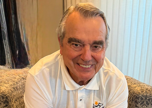 An indoor photo of Bob Zangas smiling at the camera. He has neatly combed grey hair, dark eyebrows, and is wearing a white polo shirt with a small dog logo on the left chest. He is seated on a textured patterned couch with vertical blinds in the background.