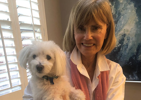 An indoor professional headshot of Diane Buhr Engelsmann smiling at the camera. She has blonde hair with bangs, is wearing a white collared shirt with a pink scarf, and is holding a small, fluffy white dog. The background shows a window with white shutters on the left and an abstract blue painting on the right.