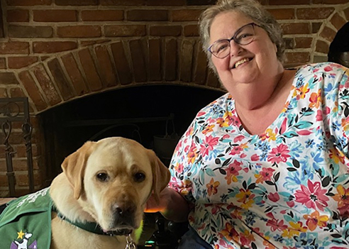 An indoor photo of Jennifer Kuehnle smiling warmly at the camera. She has short grey hair, wears eyeglasses, and is dressed in a vibrant floral-patterned blouse. She is seated next to a yellow Labrador Retriever wearing a green service vest, with a large brick fireplace in the background.