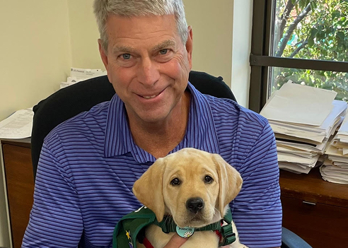 An indoor professional headshot of Jim Sietz smiling while sitting at an office desk. He has short, grey hair, blue eyes, and is wearing a purple striped polo shirt while holding a yellow Labrador Retriever puppy wearing a green service vest.