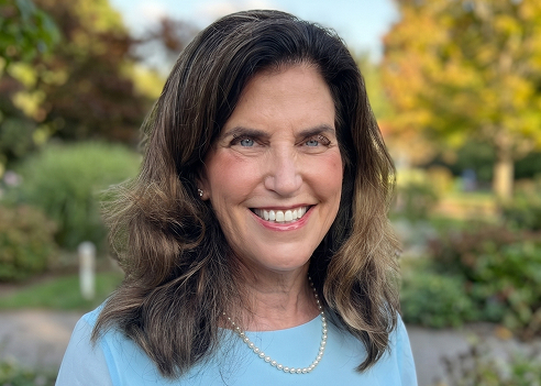 An outdoor professional headshot of a smiling woman with wavy brown hair. She has blue eyes, is wearing a light blue top with a pearl necklace, and is positioned against a softly blurred background of trees and greenery.