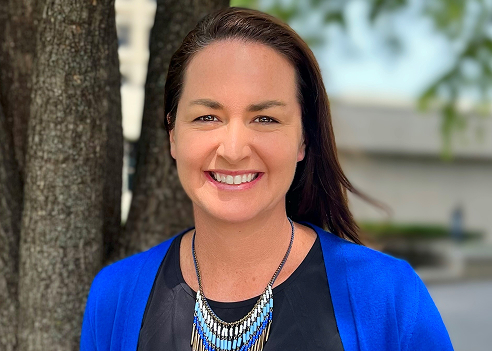 A professional outdoor headshot of a smiling woman with long brown hair pulled back. She is wearing a black top, a bright blue cardigan, and a statement necklace with blue, white, and gold beads. She is standing next to a tree trunk with a blurred outdoor background.