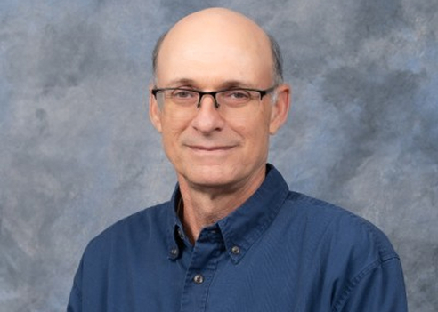 A professional headshot of Mark Anderson smiling subtly at the camera. He is a bald man wearing thin-rimmed black eyeglasses and a dark blue button-down shirt, positioned against a textured grey and blue studio backdrop.