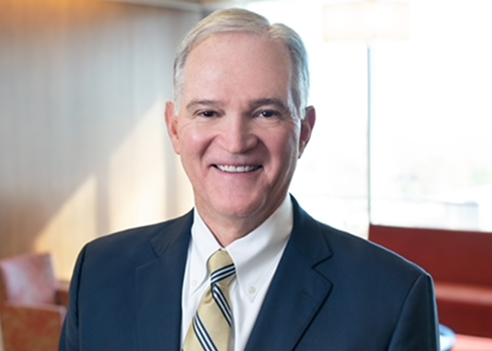 A professional indoor headshot of R. Troy Kendrick, Jr. smiling warmly at the camera. He has short, light grey-white hair, and is dressed in a dark blue suit jacket, a white collared shirt, and a gold-and-blue striped tie, set against a softly blurred office lobby background.
