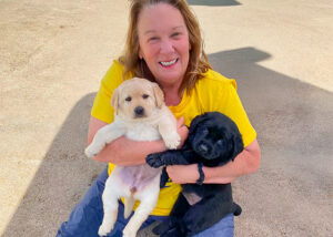 An outdoor photo of Susan Kelly smiling warmly while sitting on the ground. She has light brown hair, is wearing a bright yellow t-shirt and blue pants, and is holding two Labrador Retriever puppies—one yellow and one black—in her arms.
